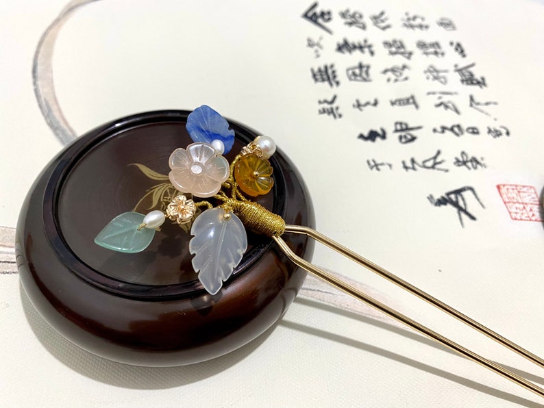 May include: A gold hair stick with a floral design. The design features a pink flower, blue and white leaves, and yellow and white beads.