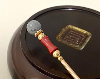 May include: A gold hair stick with a gray marble sphere on top and a red cylindrical bead in the middle.