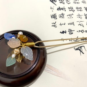 May include: A gold hair stick with a cluster of pastel colored gemstone flowers and leaves. The hair stick is resting on a dark brown wooden surface.