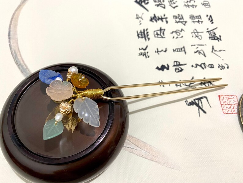 May include: A gold hair stick with a cluster of pastel colored gemstone flowers and leaves. The hair stick is resting on a dark brown wooden surface.
