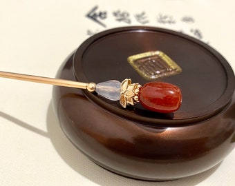May include: A gold hair stick with a white and red stone accent. The stick is resting on a dark brown wooden surface.