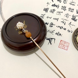 May include: A gold hair stick with a white pearl cluster, a clear crystal bead, and a brown bead. The stick is resting on a wooden surface.