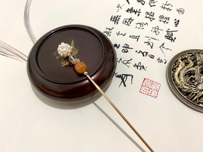 May include: A gold hair stick with a white pearl cluster, a clear crystal bead, and a brown bead. The stick is resting on a wooden surface.
