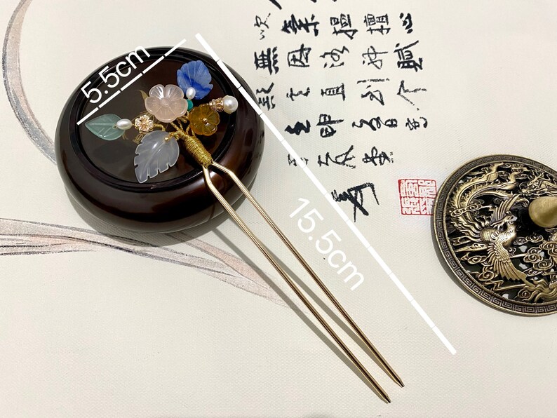 May include: A gold hair stick with a decorative floral design featuring pink, blue, green, and yellow stones. The stick is 15.5 inches long and the floral design is 5.5 inches wide.