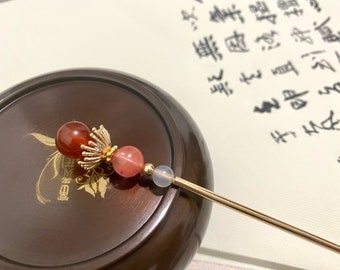 May include: A gold hair stick with three beads. The beads are red, pink, and white. The stick is on a wooden surface.