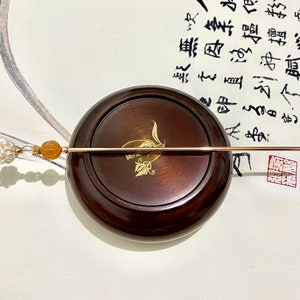 May include: A gold hair stick with a pearl and orange stone accent. The stick is resting on a wooden base.