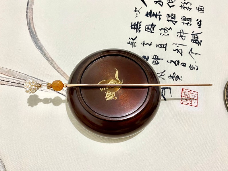 May include: A gold hair stick with a pearl and orange stone accent. The stick is resting on a wooden base.