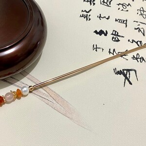 K&ouml;nnte beinhalten: Ein goldener Haarstick mit einem dekorativen Design aus orangefarbenen und wei&szlig;en Perlen. Der Stick liegt auf einer wei&szlig;en Oberfl&auml;che mit schwarzer Kalligraphie.