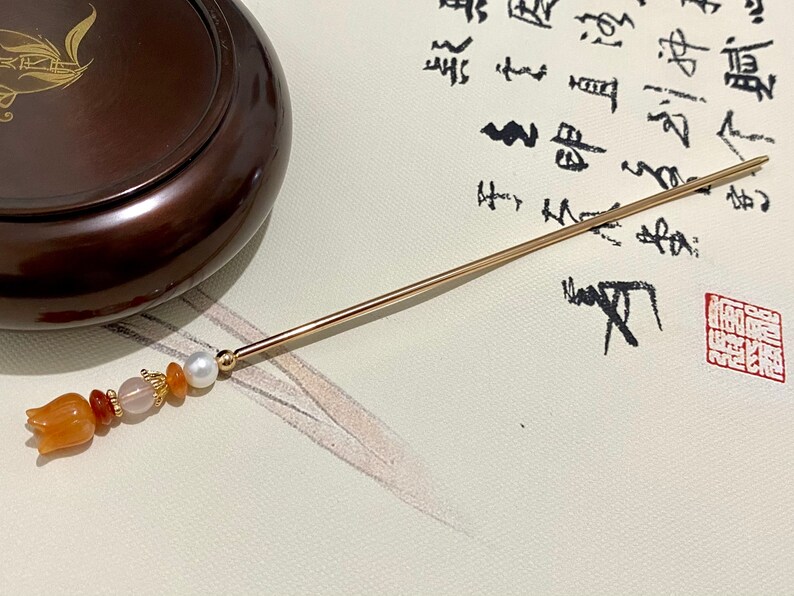 K&ouml;nnte beinhalten: Ein goldener Haarstick mit einem dekorativen Design aus orangefarbenen und wei&szlig;en Perlen. Der Stick liegt auf einer wei&szlig;en Oberfl&auml;che mit schwarzer Kalligraphie.