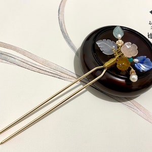 May include: A gold hair stick with a decorative floral design. The design features a variety of colored stones and beads, including blue, pink, green, and yellow. The stick is resting on a wooden base.