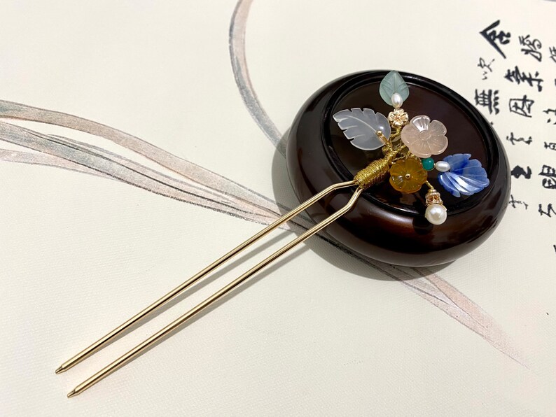 May include: A gold hair stick with a decorative floral design. The design features a variety of colored stones and beads, including blue, pink, green, and yellow. The stick is resting on a wooden base.