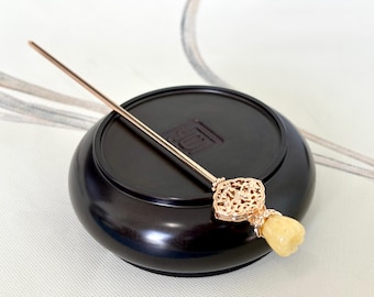 May include: A gold hair stick with a decorative, filigree design and a yellow jade stone at the end. The stick is resting on a black wooden base.