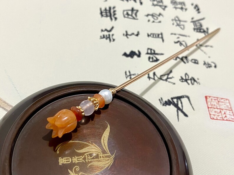 K&ouml;nnte beinhalten: Ein goldener Haarstecker mit einer geschnitzten orangefarbenen Blume und Perlen in Orange-, Rosa- und Wei&szlig;t&ouml;nen. Der Stecker liegt auf einer dunkelbraunen Holzschale.