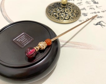 May include: A gold hair stick with three beads. The beads are red, orange, and gold. The stick is resting on a dark brown wooden surface.