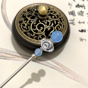May include: A silver hair stick with three light blue beads and a silver flower charm. The stick is resting on a black and gold surface.