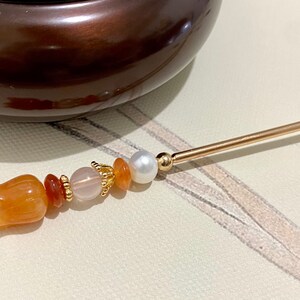 K&ouml;nnte beinhalten: Ein goldener Haarstecker mit orangefarbenen und rosafarbenen Perlen, einer wei&szlig;en Perle und einer geschnitzten orangefarbenen Blume am Ende.