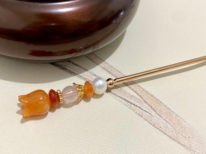 K&ouml;nnte beinhalten: Ein goldener Haarstecker mit orangefarbenen und rosafarbenen Perlen, einer wei&szlig;en Perle und einer geschnitzten orangefarbenen Blume am Ende.