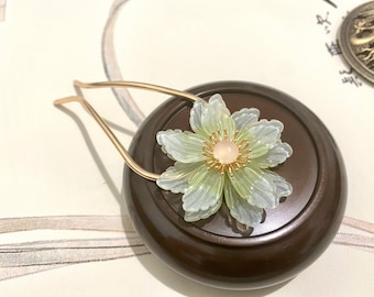 Puede incluir: Una pinza para el pelo dorada con un delicado diseño de flor verde y blanca. La flor tiene un centro rosa y detalles dorados.