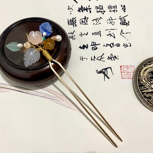 May include: A gold hair stick with a decorative top featuring a cluster of pastel pink, blue, and green flowers and leaves. The stick is resting on a dark brown wooden base.