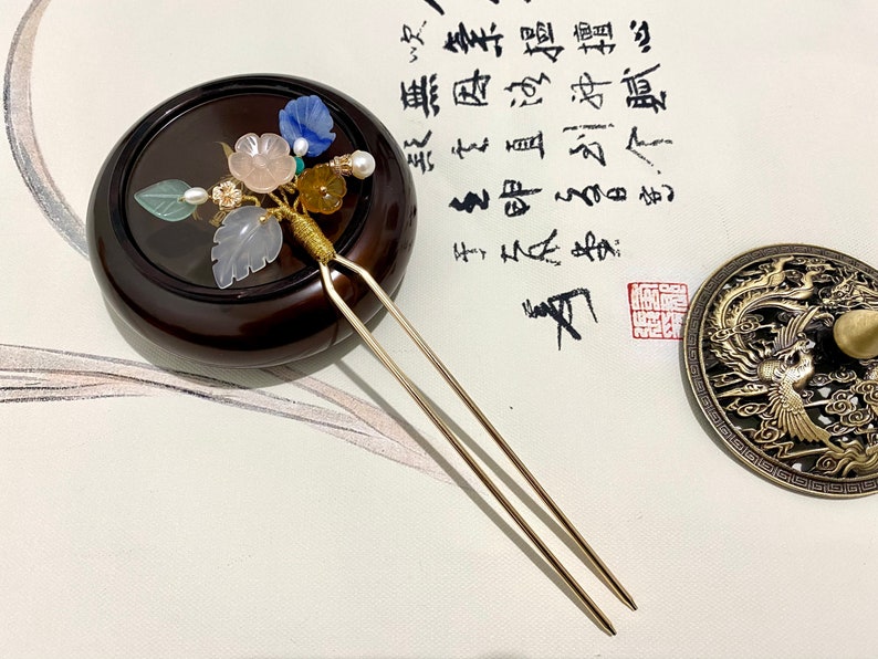 May include: A gold hair stick with a decorative top featuring a cluster of pastel pink, blue, and green flowers and leaves. The stick is resting on a dark brown wooden base.