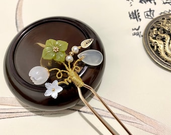 May include: A gold hair stick with a delicate design featuring green and white flowers, pearls, and a white stone. The stick is resting on a dark brown wooden surface.