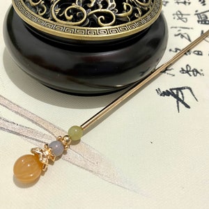 Op de afbeelding: Een gouden en oranje kralen haarstok met een decoratieve vlinderbedel. De stok ligt op een wit oppervlak met zwarte kalligrafie.