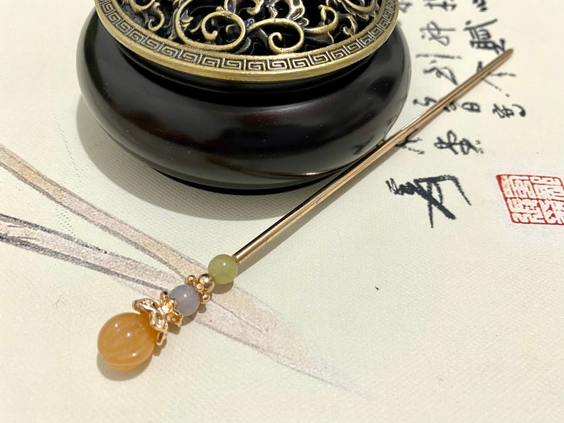Op de afbeelding: Een gouden en oranje kralen haarstok met een decoratieve vlinderbedel. De stok ligt op een wit oppervlak met zwarte kalligrafie.