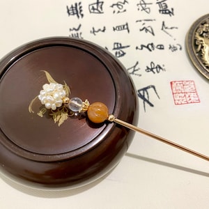 May include: A gold hair stick with a white pearl cluster, a clear crystal bead, and an orange agate bead. The stick is resting on a wooden box with a gold design.