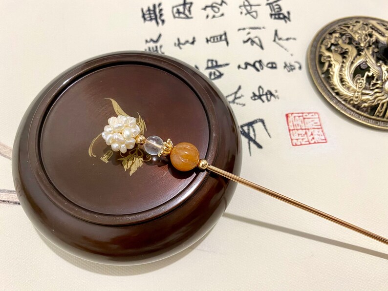 May include: A gold hair stick with a white pearl cluster, a clear crystal bead, and an orange agate bead. The stick is resting on a wooden box with a gold design.