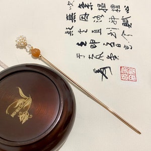 May include: A gold hair stick with a pearl and orange stone accent. The stick is resting on a wooden box with a gold design. The box is sitting on a white surface with Chinese calligraphy.