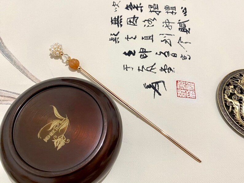 May include: A gold hair stick with a pearl and orange stone accent. The stick is resting on a wooden box with a gold design. The box is sitting on a white surface with Chinese calligraphy.