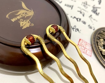 May include: Two gold-colored hairpins with a wavy design. Each hairpin features a red-brown bead at the top and is accented with wire wrapping. The background includes a dark brown wooden container and calligraphy.