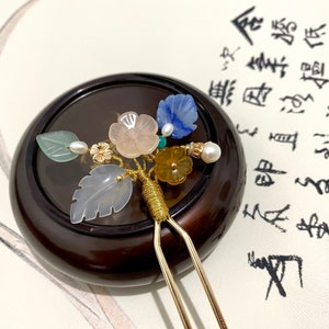 May include: A gold hair stick with a decorative floral design. The design features a variety of colorful stones, including green, blue, pink, and yellow. The hair stick is resting on a wooden surface.