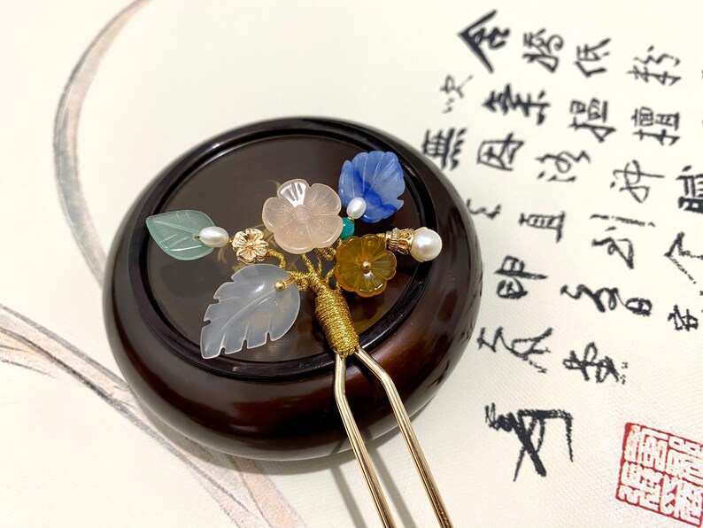 May include: A gold hair stick with a decorative floral design. The design features a variety of colorful stones, including green, blue, pink, and yellow. The hair stick is resting on a wooden surface.