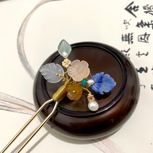 May include: A gold hair stick with a decorative cluster of jade, blue, and pink stones, leaves, and pearls. The hair stick is resting on a dark wood surface.