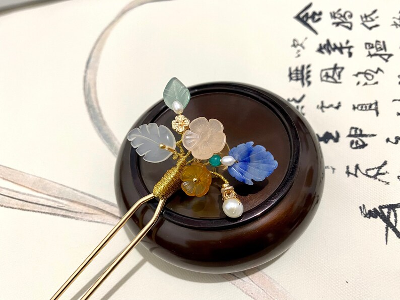 May include: A gold hair stick with a decorative cluster of jade, blue, and pink stones, leaves, and pearls. The hair stick is resting on a dark wood surface.