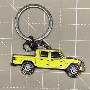 May include: A yellow metal keychain featuring a detailed model of a Jeep Gladiator pickup truck. The truck has black wheels and a silver metal ring.