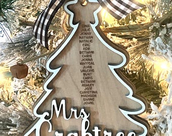 May include: A wooden Christmas tree ornament with a white outline and the text "Mrs. Crabtree 2024". The ornament is decorated with a black and white checkered ribbon and has a list of names on the front.