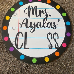May include: A round, black-framed sign with a white background and colorful polka dots around the edge. The sign says "Mrs. Ayla's Class" in black cursive writing.