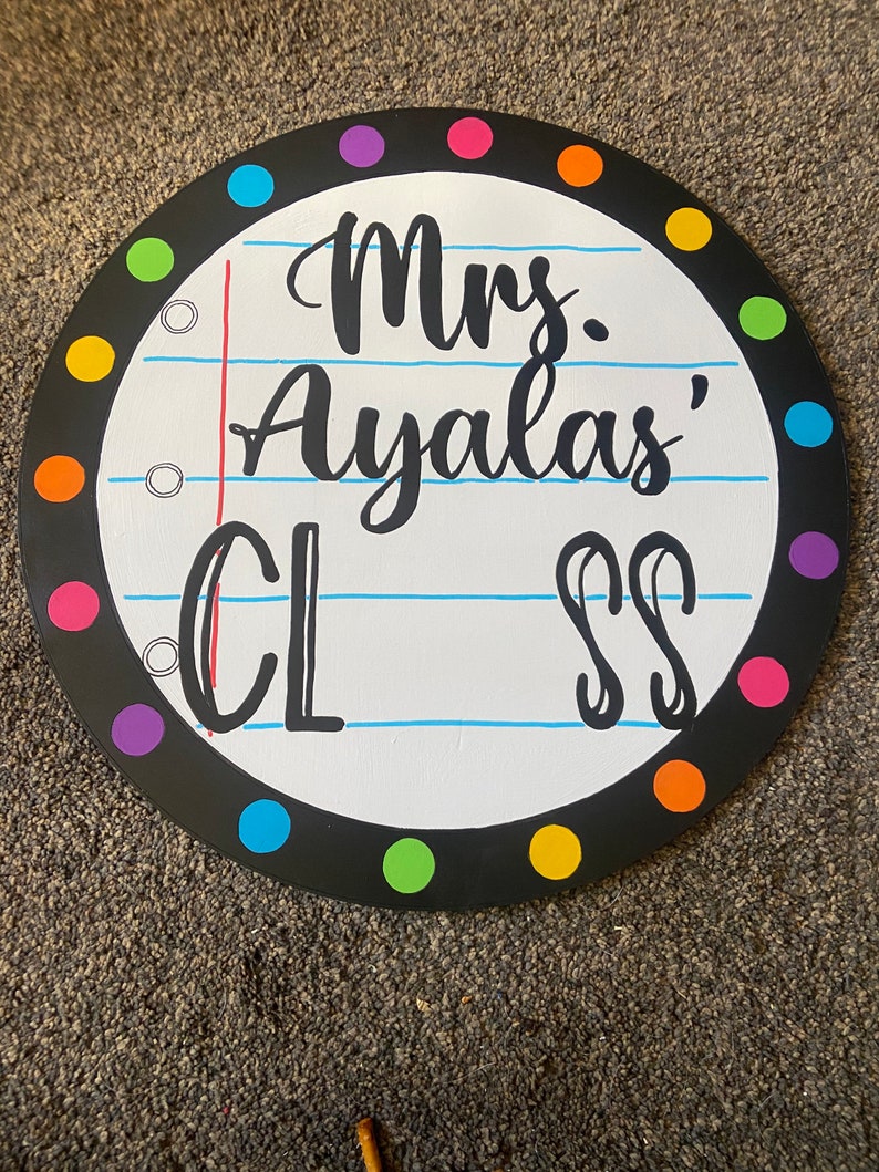 May include: A round, black-framed sign with a white background and colorful polka dots around the edge. The sign says "Mrs. Ayla's Class" in black cursive writing.