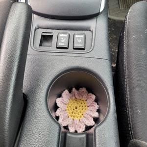 May include: A pink and yellow crocheted flower coaster sits in a cup holder in a car. The cup holder is made of black plastic and is located in the center console of the car.
