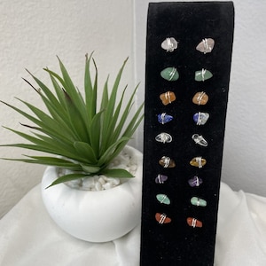 May include: A display of 12 pairs of gemstone earrings. Each pair features a different colored stone, including pink, green, orange, blue, clear, and brown. The stones are wrapped in silver wire and attached to silver posts.