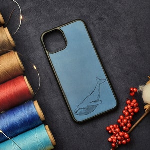 Może przedstawiać: Niebieskie skórzane etui na telefon z wzorem wieloryba. Etui leży na ciemnoszarym tle z szpulami nici w różnych kolorach.