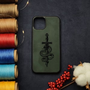 Puede incluir: Funda de teléfono verde oliva con un diseño de serpiente y espada en negro. El diseño presenta una serpiente enroscada alrededor de una espada. La funda tiene un borde negro y un fondo oscuro. Accesorio.