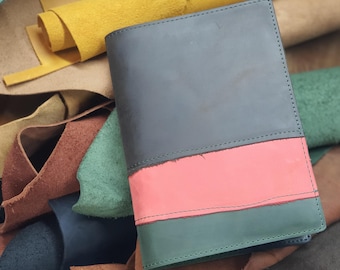 May include: A black leather notebook with a pink and green leather cover. The notebook is surrounded by pieces of leather in various colors.
