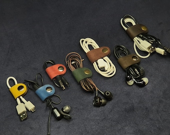 Op de afbeelding: Assortiment van zeven leren snoerorganisatoren in verschillende kleuren, elk met een metalen knoopsluiting. De organisatoren houden verschillende soorten snoeren vast, waaronder koptelefoonsnoeren en oplaadkabels.