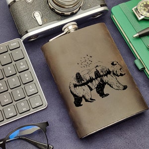 Pode incluir: Uma garrafa de couro marrom com um desenho gravado em preto de um urso caminhando por uma floresta com pássaros voando acima.