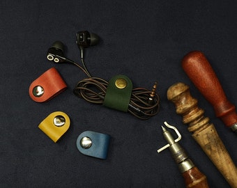 May include: Four leather cord organizers in red, yellow, blue, and green with metal accents. The organizers are on a dark blue surface with a pair of black earbuds and two leather working tools.