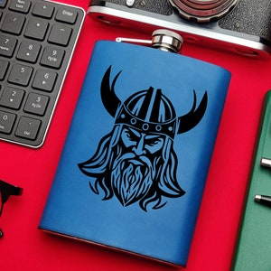 Puede incluir: Una petaca de acero inoxidable azul con un diseño de guerrero vikingo negro. El guerrero lleva un casco con cuernos y tiene barba.
