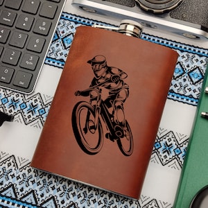 May include: A brown leather flask with a black silhouette of a mountain biker riding a bike. The flask has a silver top.